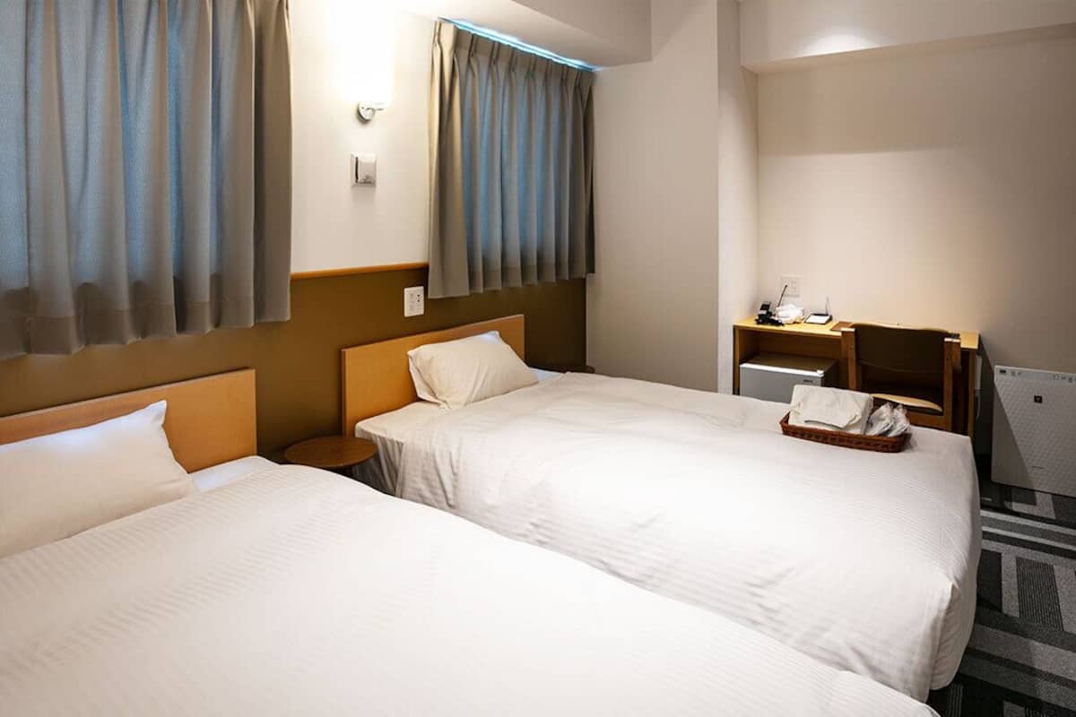 A hotel room with two beds, a desk, a chair, and a small table with a basket.