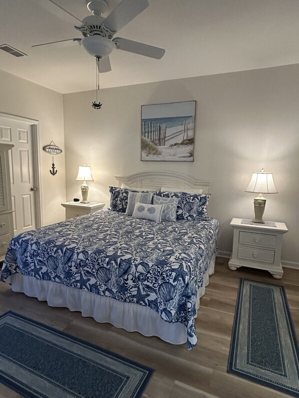 A bedroom with a bed, two bedside tables, a ceiling fan, a welcome sign, a beach-themed painting, and a rug.