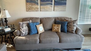 Een woonkamer met een beige bank, gestreepte kussens, een glazen salontafel en een groot abstract schilderij aan de muur.