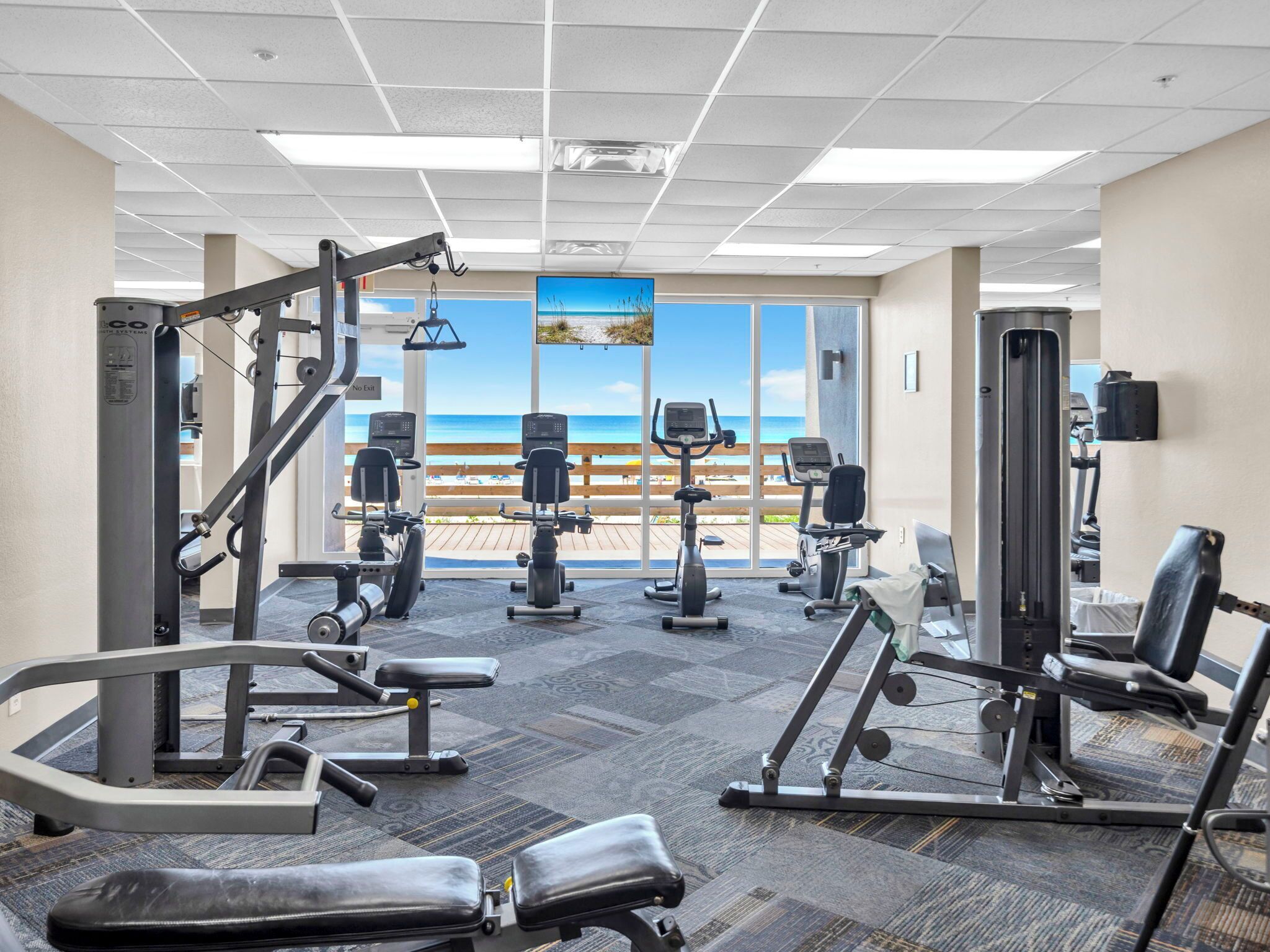 Un gimnasio con diversos equipos de ejercicio, ventanas amplias con vista al mar e iluminación empotrada en el techo.