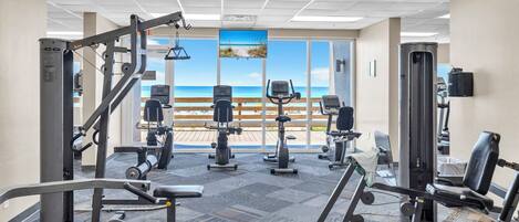 Un gimnasio con diversos equipos de ejercicio, ventanas amplias con vista al mar e iluminación empotrada en el techo.