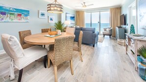 A spacious living area with a round wooden dining table, wicker chairs, a gray sofa, and a ceiling fan.