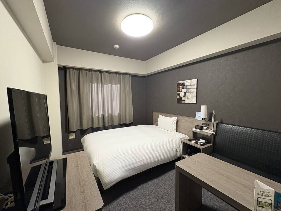 A hotel room with a bed, a desk, a chair, a TV, a bedside table, a lamp, and a wall-mounted artwork.