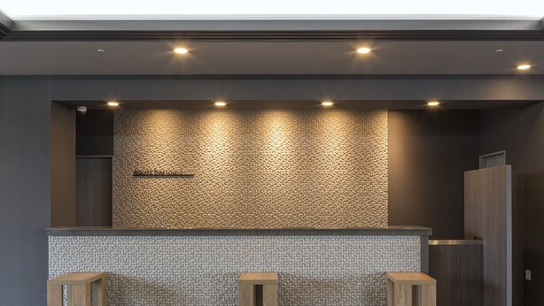A reception area with a textured wall, a reception desk, and three wooden stools. - Hotel Route Inn Shinfuji Ekiminami (Fuji)