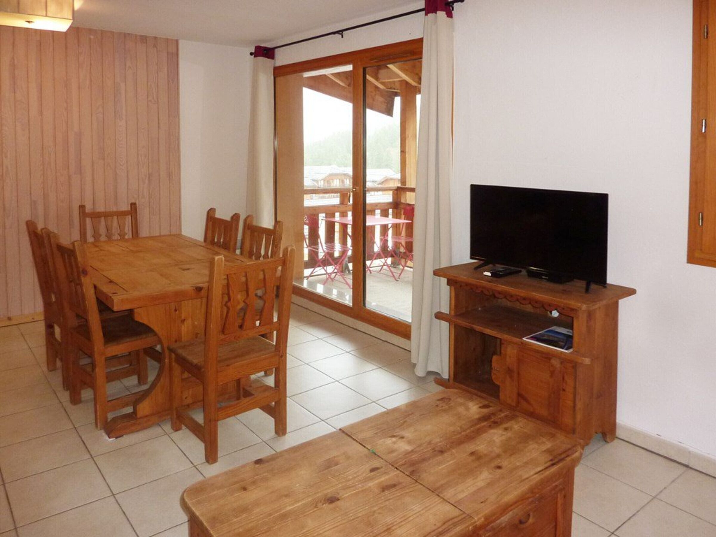Une table à manger en bois, des chaises, un banc, un téléviseur posé sur un meuble en bois, et une porte-fenêtre coulissante donnant sur un espace extérieur équipé de chaises.