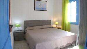 A bedroom with a bed, two bedside tables with lamps, a framed picture on the wall, and a window with curtains. - Ground floor, 5 places (Costa Rei)