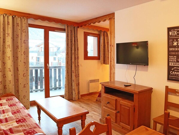 Una habitación de hotel con una mesa de madera, sillas, un televisor y un balcón con cortinas.