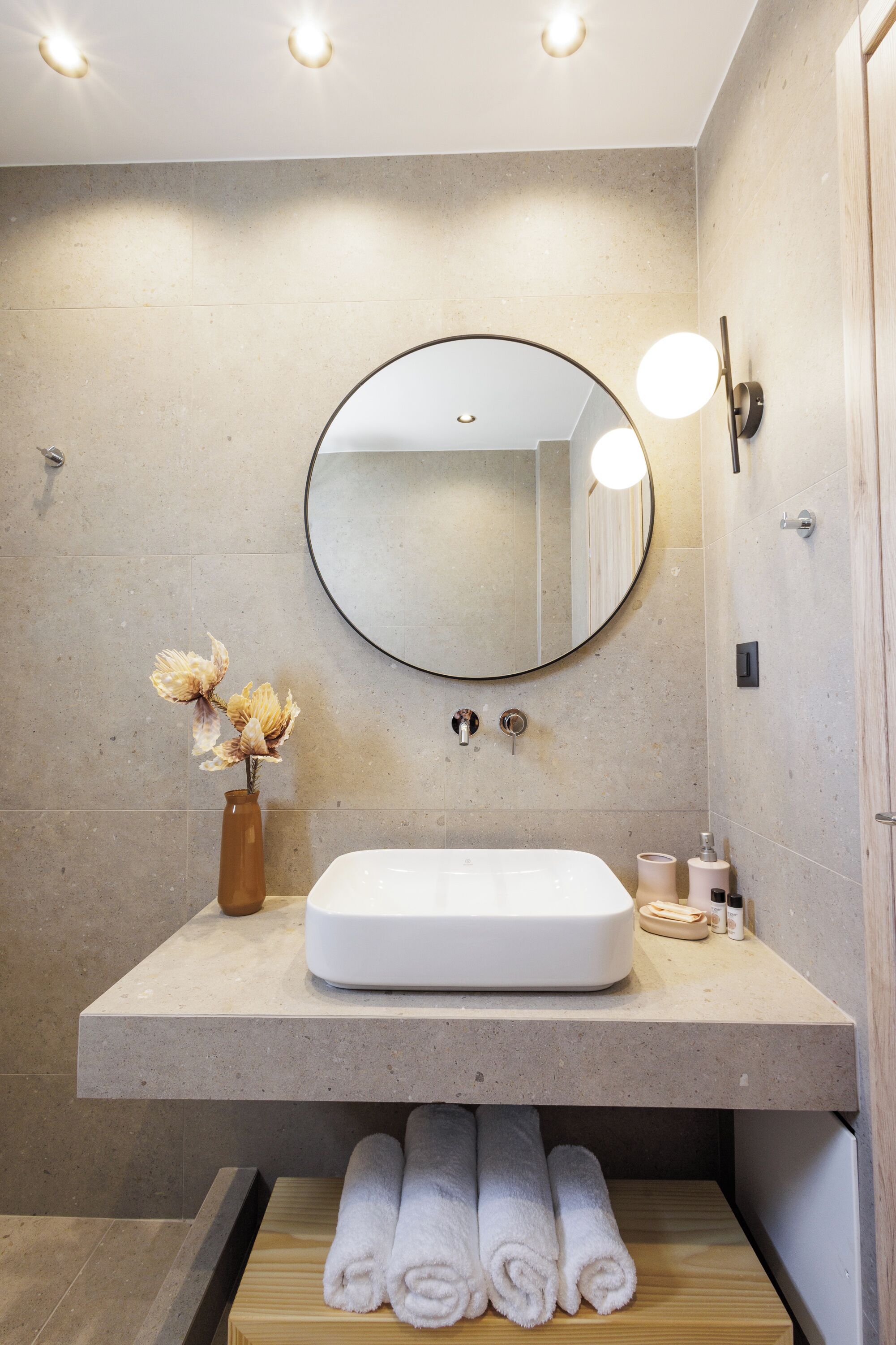 Un baño con espejo redondo, lavabo blanco y un jarrón con flores secas.