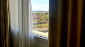Una ventana con cortinas entreabiertas, mostrando una vista de paisaje rural con campos y edificios lejanos.