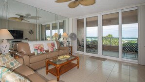 A spacious living room with a large sofa, a coffee table, and a television. There is a balcony with a view of the ocean. - Crescent Beach Club 3B Beachfront Views of the Gulf (Clearwater Beach)