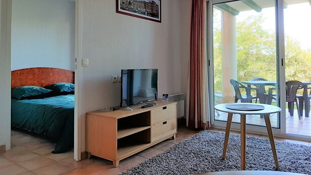 Quarto com cama, televisão sobre um suporte de madeira, uma pequena mesa e varanda com cadeiras.