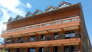 Un edificio de varios pisos con balcones, un letrero que indica 'Kamusi Kings Guest House' ofreciendo alojamiento las 24 horas, y un coche estacionado al frente.
