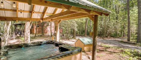 A hot tub under a wooden pergola in a forested area with a rustic cabin in the background.