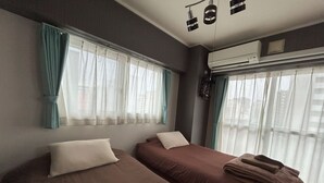 Ein Zimmer mit zwei Betten, jeweils mit Kissen und Decke. Eine Klimaanlage ist an der Wand angebracht und ein Fenster mit Vorhängen vorhanden.