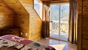 Ein gemütliches Blockhauszimmer mit Holzbalkendecke, einem großen Fenster mit Vorhängen, einem Bett mit Quilten und einem kleinen Nachttisch.