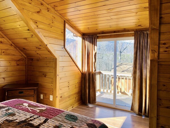 Relax in this cabin-style bedroom with a sloped ceiling, featuring a large window and a comfortable quilted bedspread.