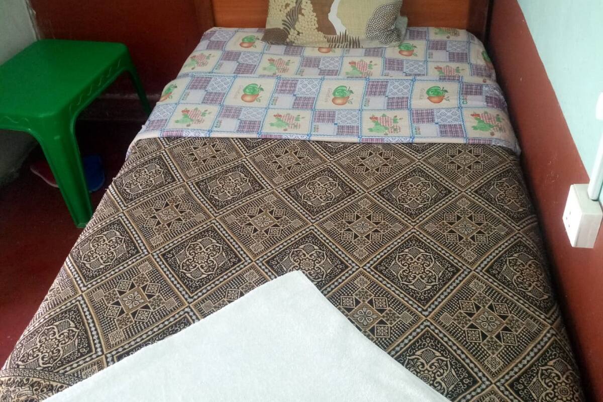 Un lit simple avec une couvre-lit à motifs et une couverture blanche. Un tabouret en plastique vert est visible sur le côté gauche du lit.