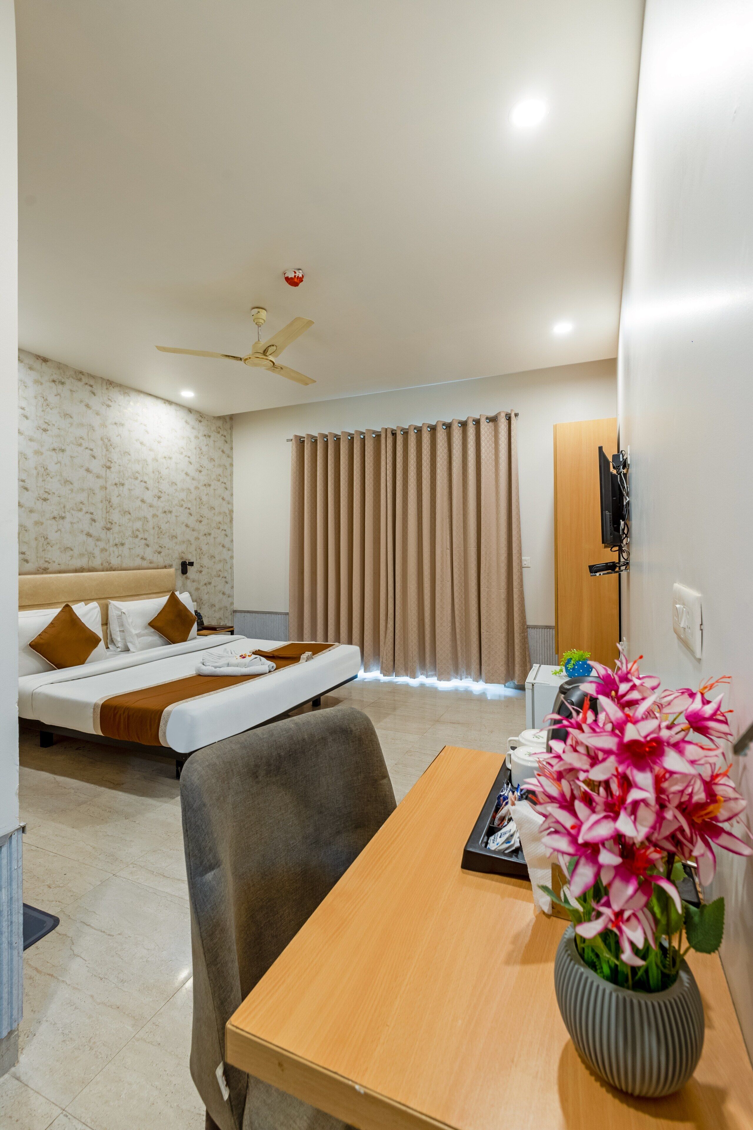 Une chambre d’hôtel équipée d’un lit, d’un bureau, d’une chaise, d’un ventilateur de plafond, d’un téléviseur fixé au mur et d’une fenêtre avec des rideaux.