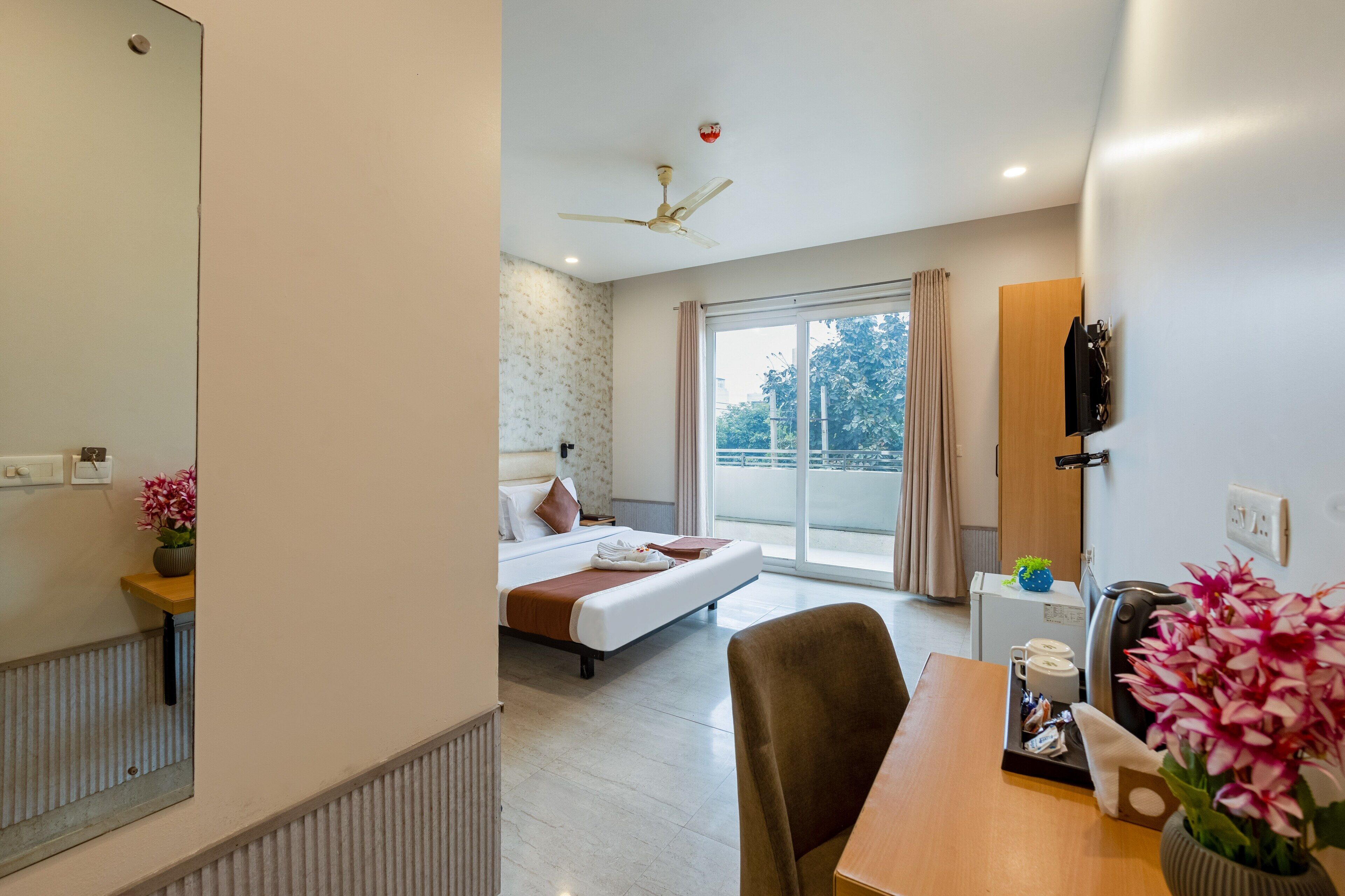 Habitación de hotel moderna con cama, escritorio, silla, ventilador de techo y un amplio ventanal con vistas a árboles.