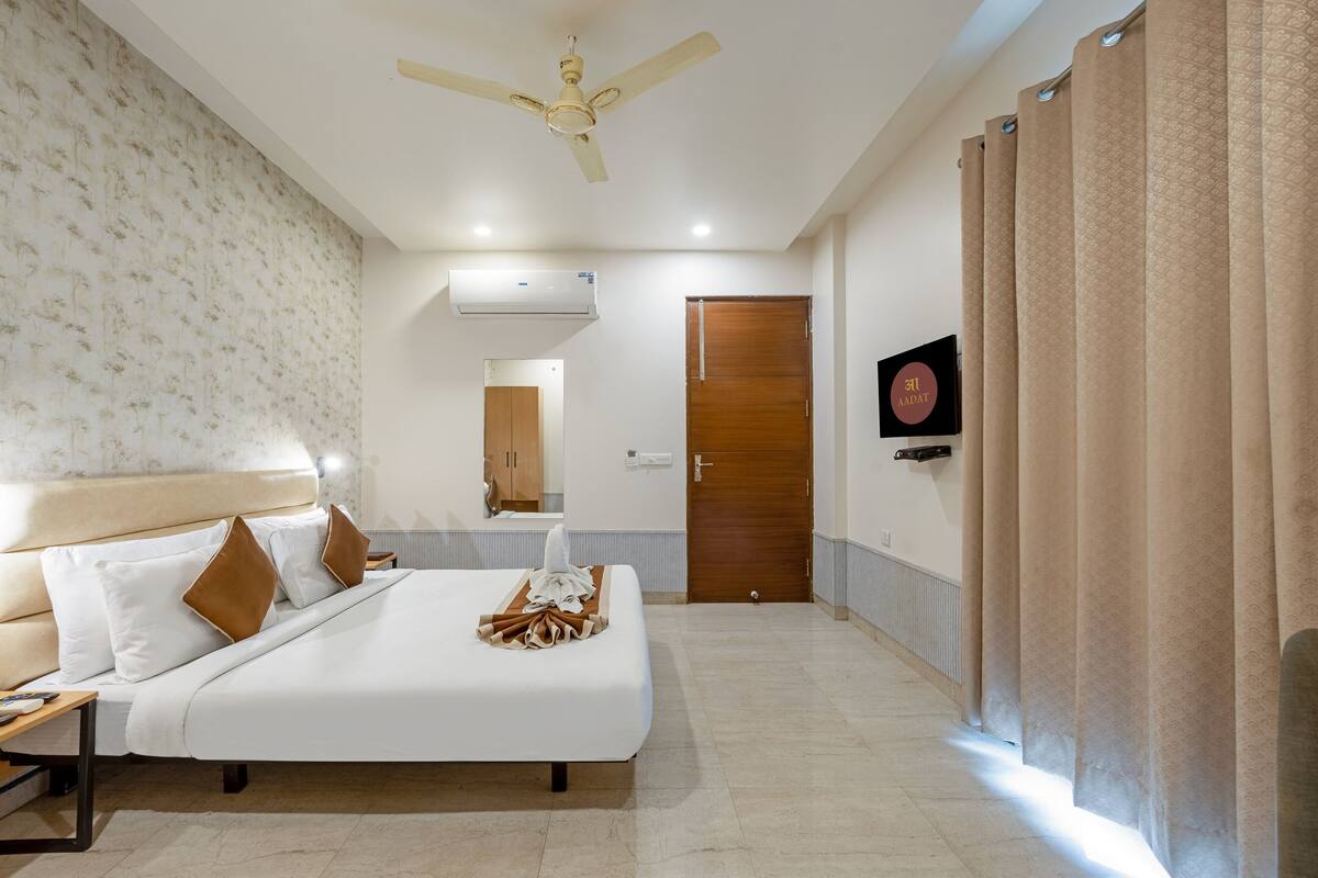 A hotel room with a bed, a ceiling fan, an air conditioning unit, a wooden door, and a television on the wall.