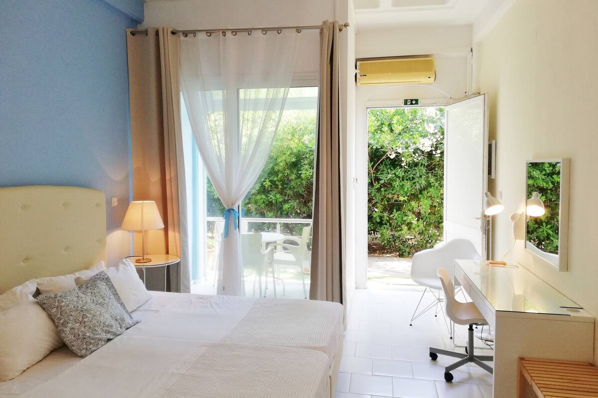 A hotel room with a bed, a desk, a chair, and a view of greenery outside.