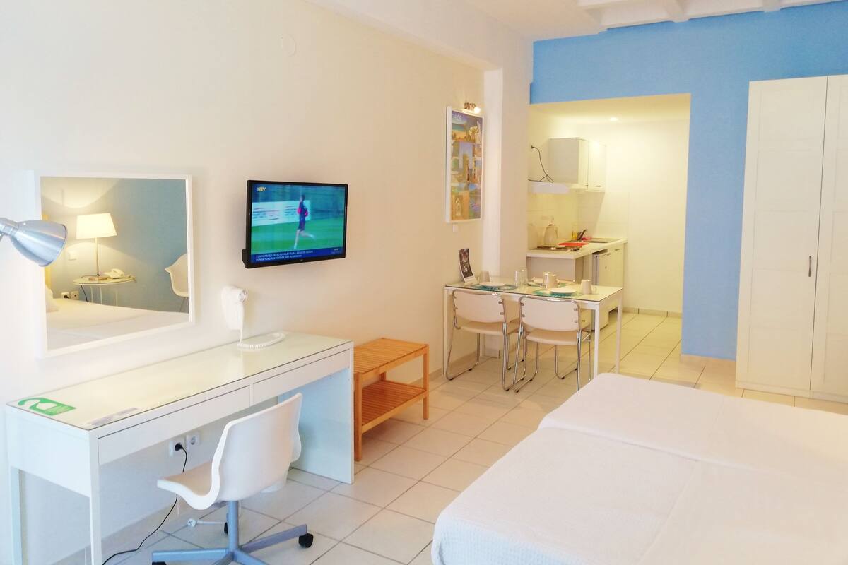 A hotel room with a bed, a desk with a chair, a TV, a small table with a chair, a sink, and a wardrobe.