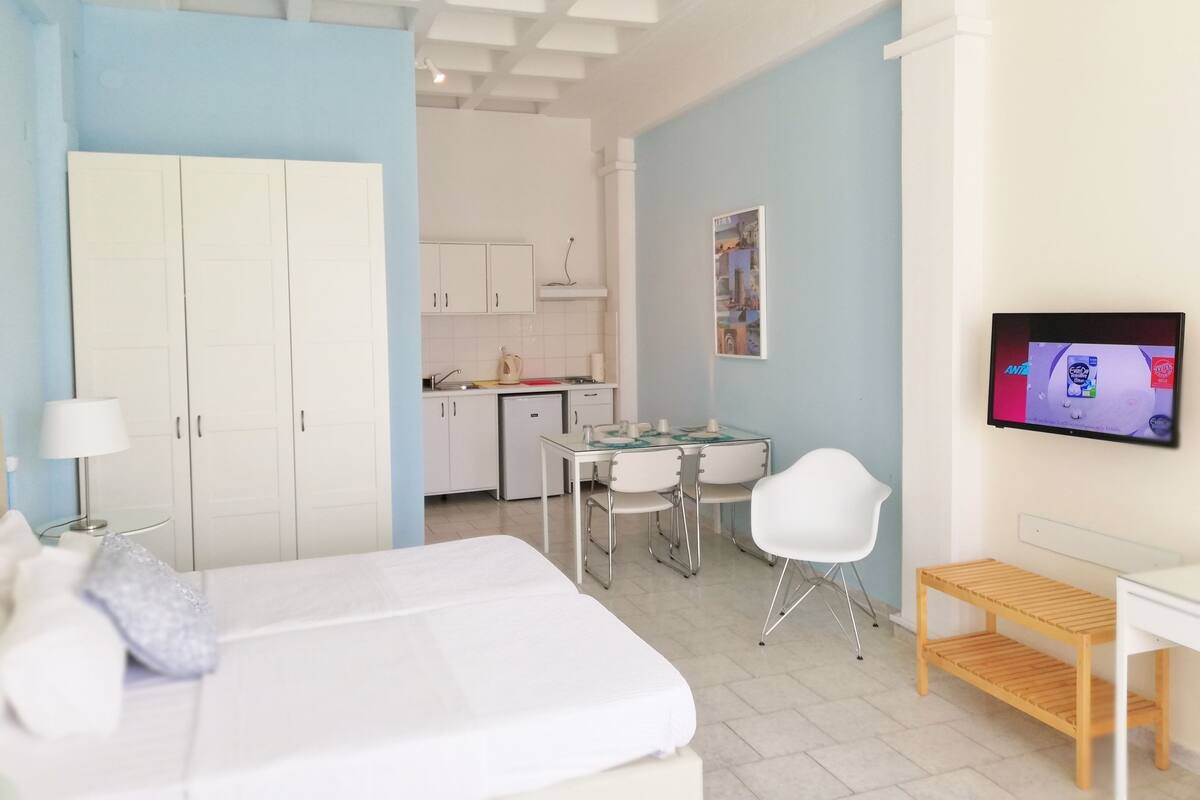 A modern hotel room with a bed, a desk, a chair, a TV, and a kitchenette.