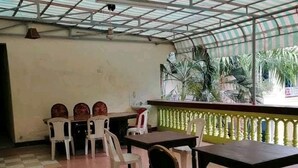 Un espacio al aire libre techado con una mesa y sillas, un ventilador de techo y vistas a palmeras a través de una barrera de vidrio.