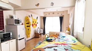 A bedroom with a bed featuring a colorful Pokémon-themed duvet, a small desk with a computer and a fan, a microwave, and a refrigerator. - PEOPLE NoIII 810 (Sapporo)