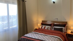 Ein Hotelzimmer mit einem Bett, einem Nachttisch, einer Lampe, einem Fenster mit Vorhängen und einer Wandleuchte.