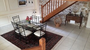 Un coin repas avec une table en verre entourée de chaises, un escalier en bois, un mur en pierre et un petit meuble en bois.