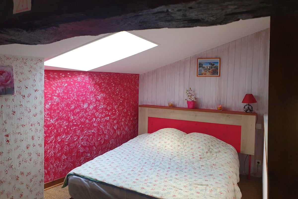 Een slaapkamer met een schuine kap, een dakraam, een bed met bloemenbeddengoed, een rood hoofdbord, een klein nachtkastje met een lamp, en een ingelijst schilderij aan de muur.