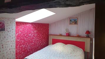 A bedroom with a sloped ceiling, a skylight, a bed with floral bedding, a red headboard, a small bedside table with a lamp, and a framed picture on the wall.