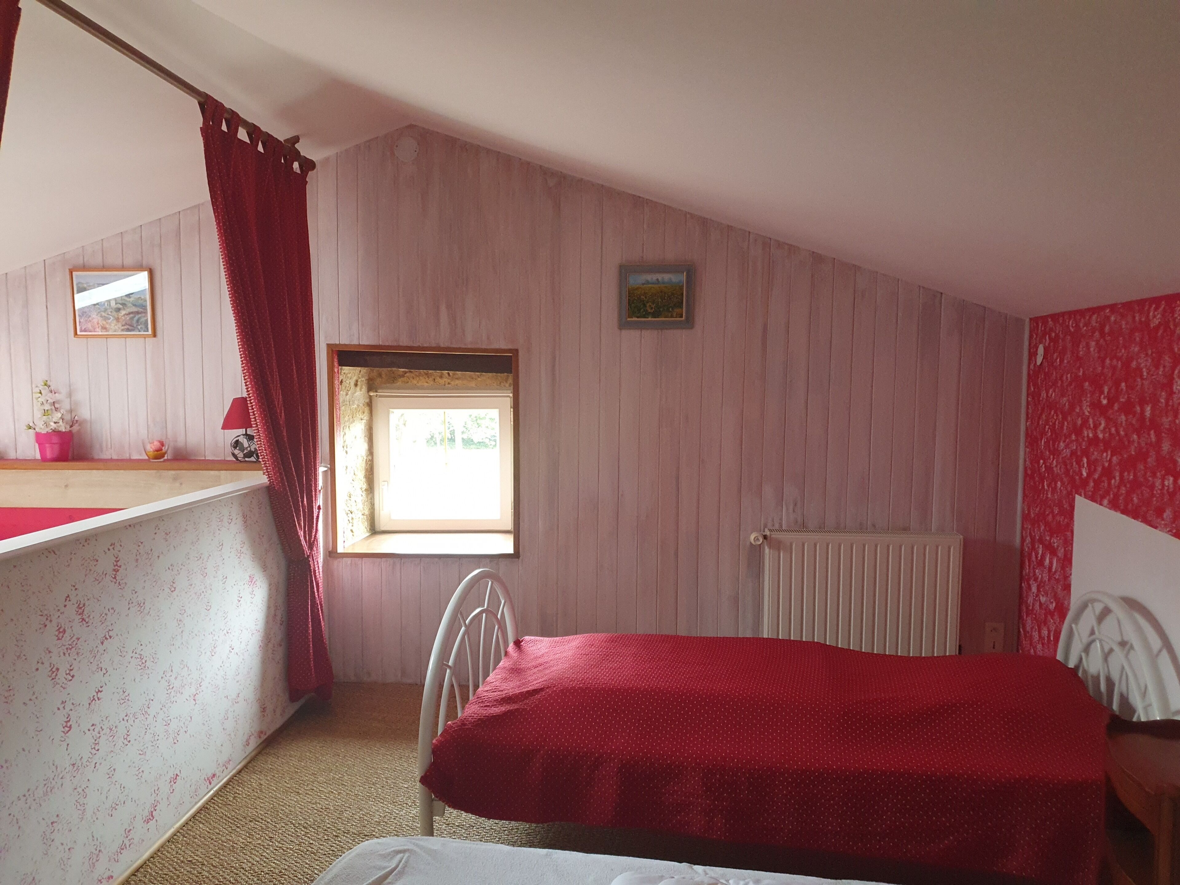 A bedroom with a sloped ceiling, a window, a radiator, a bed with a red cover, a chair, and a small table with a pink flower vase.