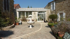 Une cour intérieure agrémentée d’une fontaine, de plantes en pot et d’un bâtiment doté de grandes fenêtres et d’une façade en pierre.