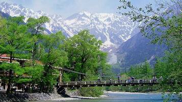 A wooden bridge over a river with people walking, surrounded by trees and snow-capped mountains in the background.