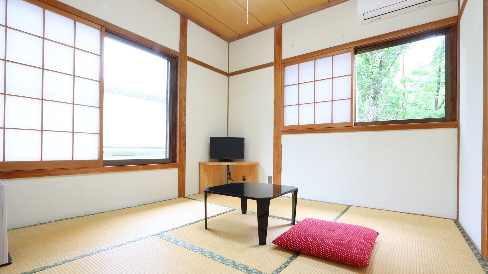 A traditional Japanese room with tatami flooring, a black table, a red cushion, and a television on a wooden stand.