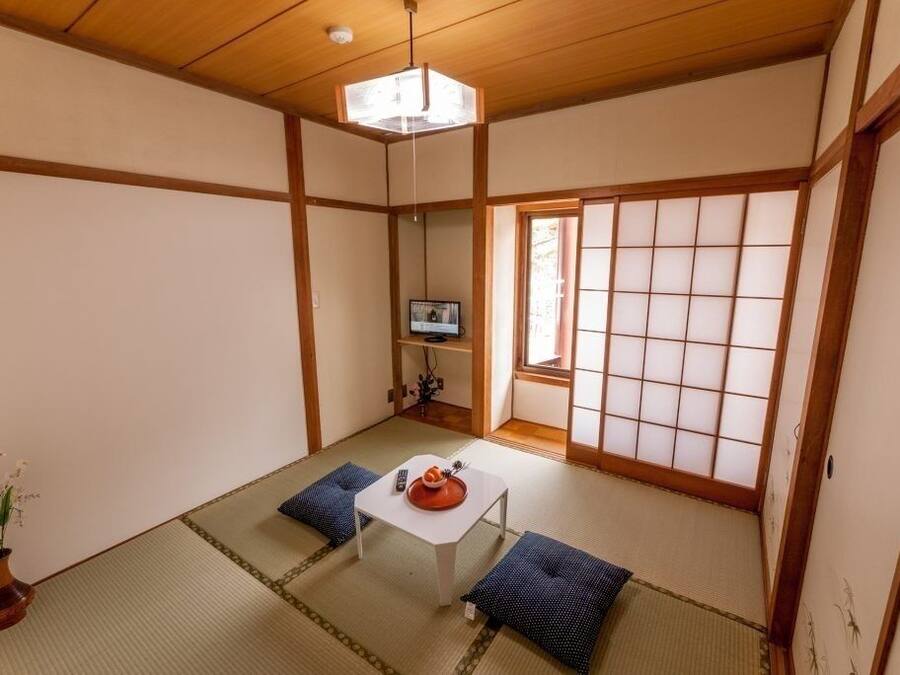 Um quarto japonês tradicional com piso de tatame, uma mesa baixa com almofadas, uma televisão em uma prateleira e uma porta de papel shoji.