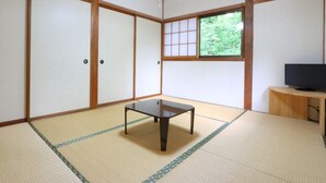 A traditional Japanese room with tatami flooring, sliding doors, a small table, a TV on a wooden stand, and a window with a view of greenery. - kongoji syukubo seihoin (Azumino)