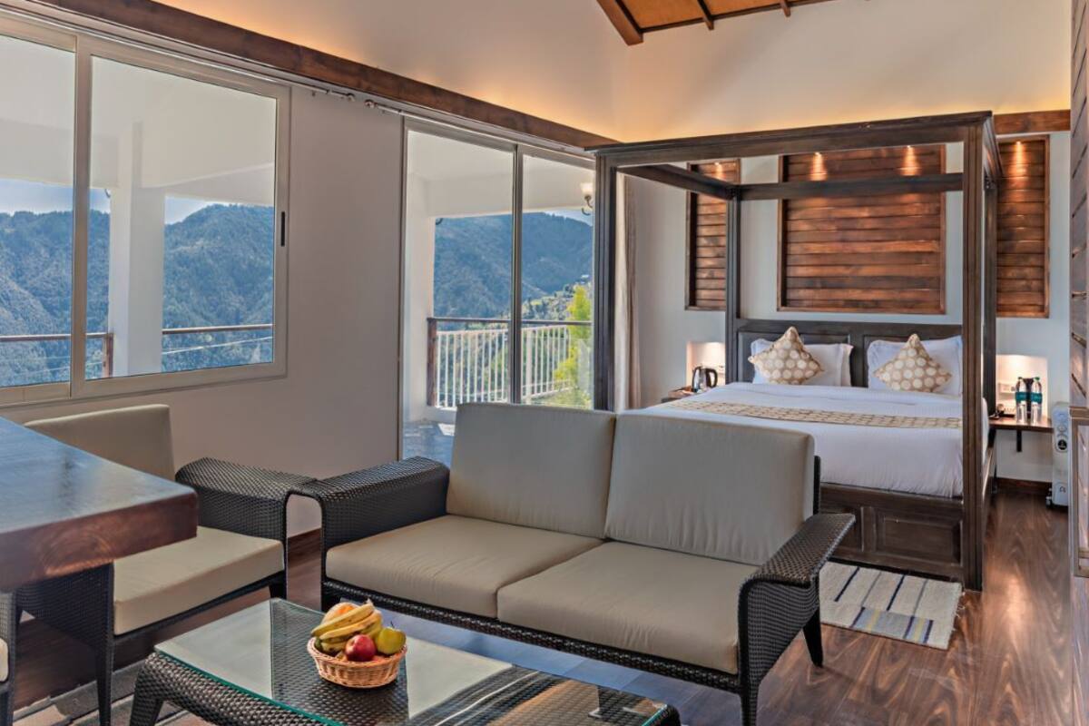 A modern hotel room with a large bed, a balcony with a mountain view, wicker furniture, and a glass table with fruit.