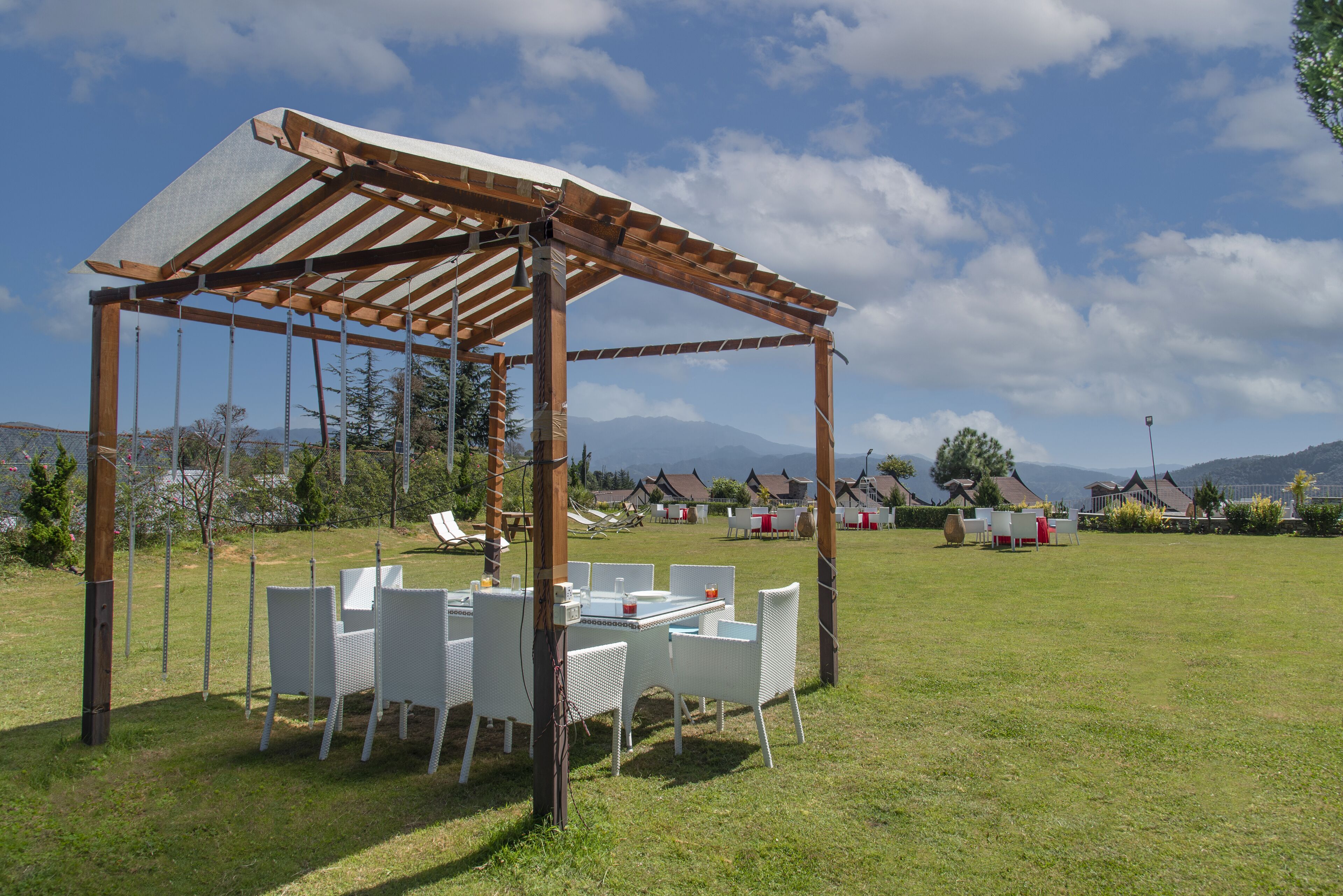 Un espacio de estar al aire libre cubierto con sillas blancas y una mesa, rodeado por un campo verde y un cielo despejado.