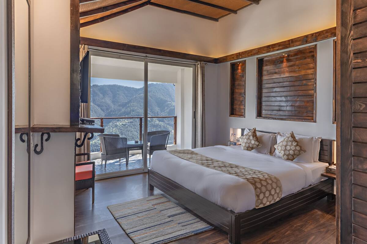 A bedroom with a large bed, a balcony with a view of the mountains, and a wooden ceiling.