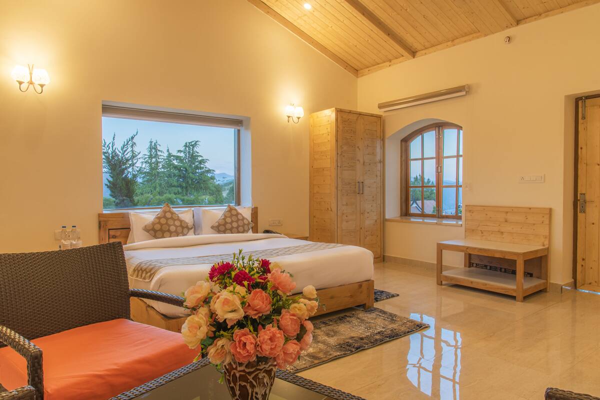 A bedroom with a bed, a wooden bench, a wicker chair, a vase of flowers, and a large window with a view of trees.