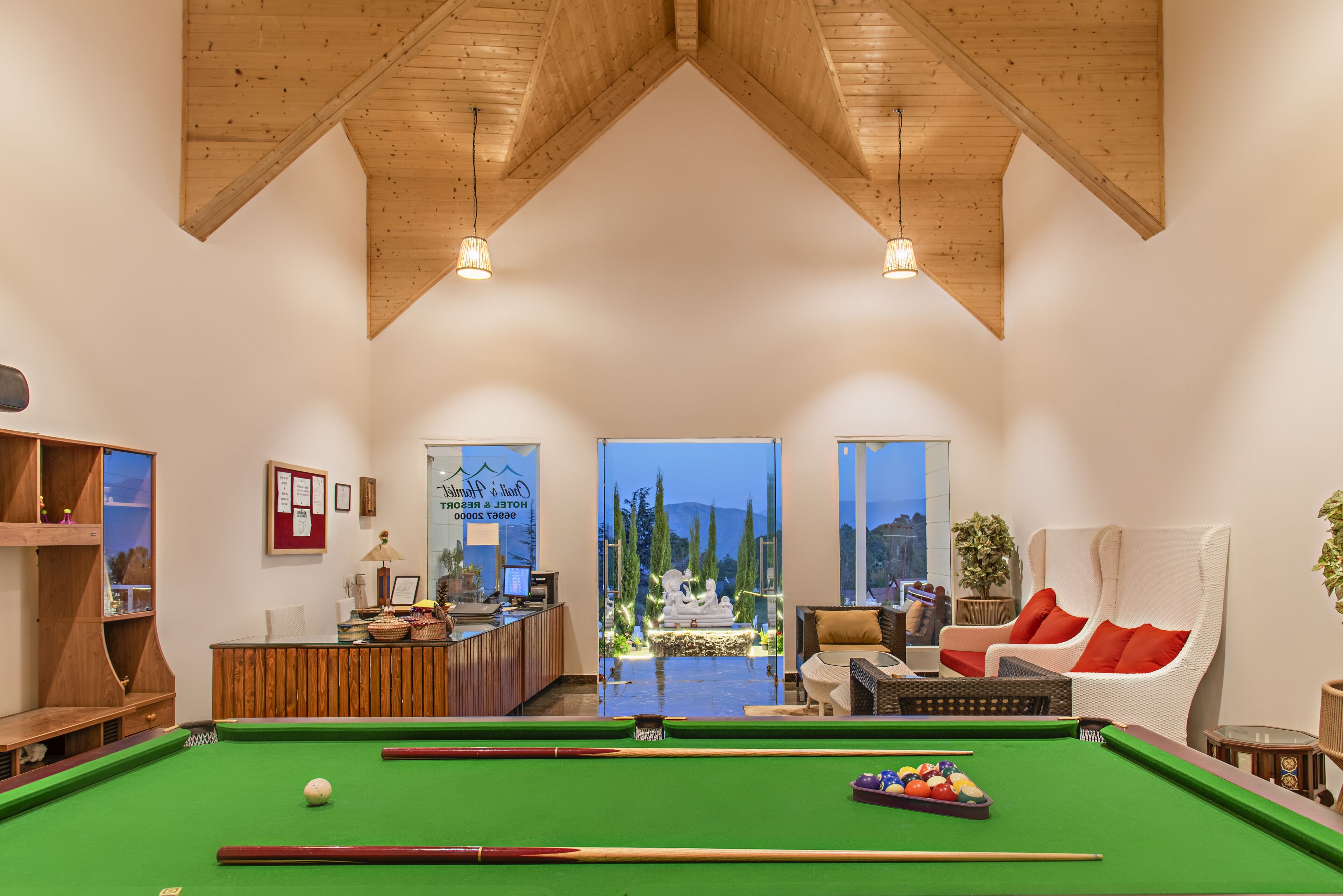 Een pooltafel in een ruimte met houten balken, een receptiebalie en uitzicht op bomen door glazen deuren.
