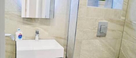 A modern bathroom with a white sink, dark cabinet, and a toilet. There are shelves with plants and a light fixture above the sink.