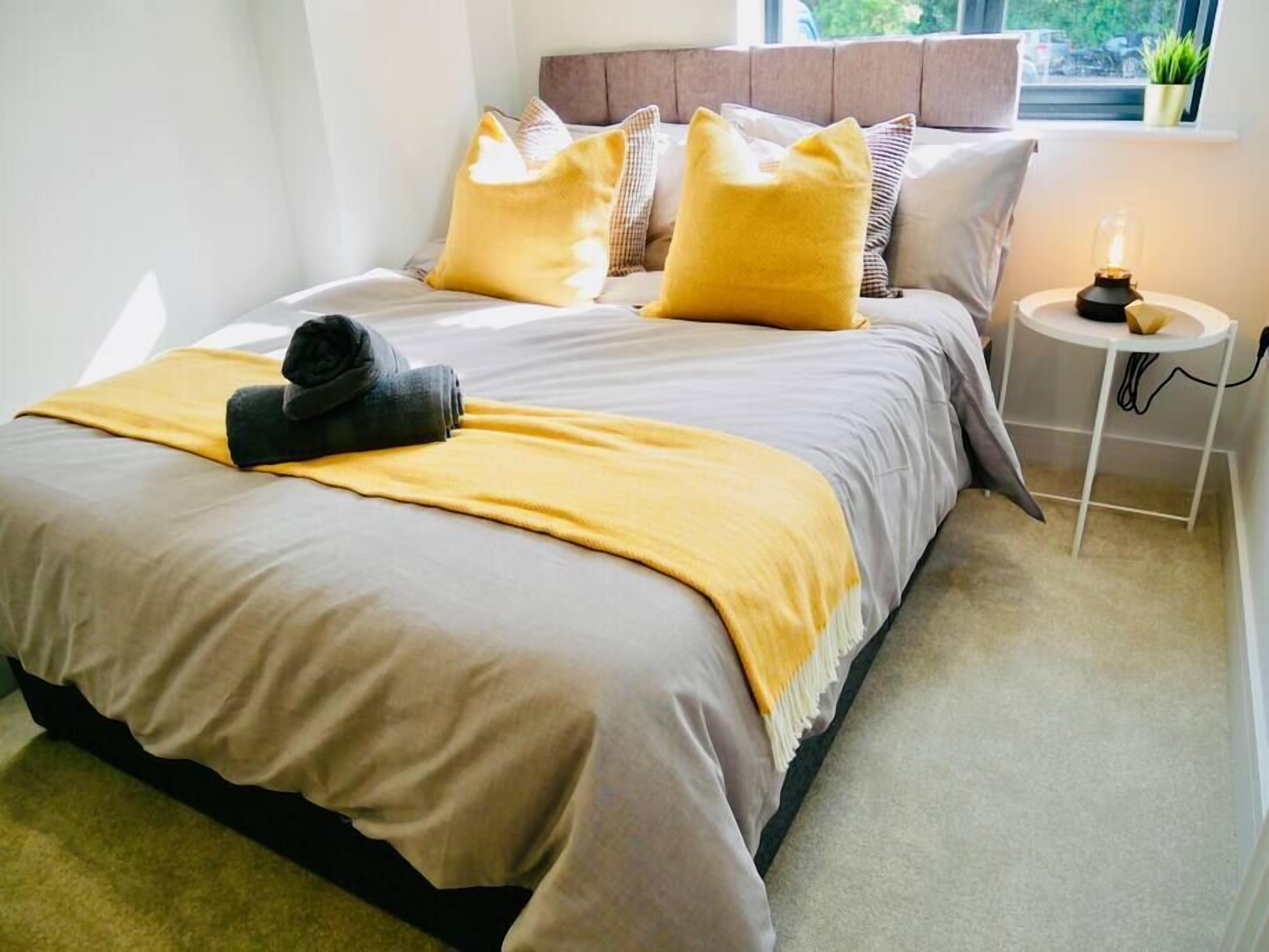 Una cama bien tendida con un edredón amarillo y una toalla enrollada, una mesita de noche con una lámpara, y una ventana con vistas a zonas verdes.