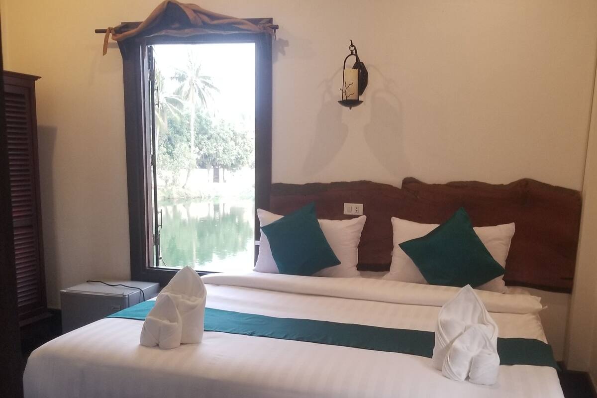 A neatly made bed with white linens and two green pillows, a bedside table with a lamp, and a window with a view of greenery.