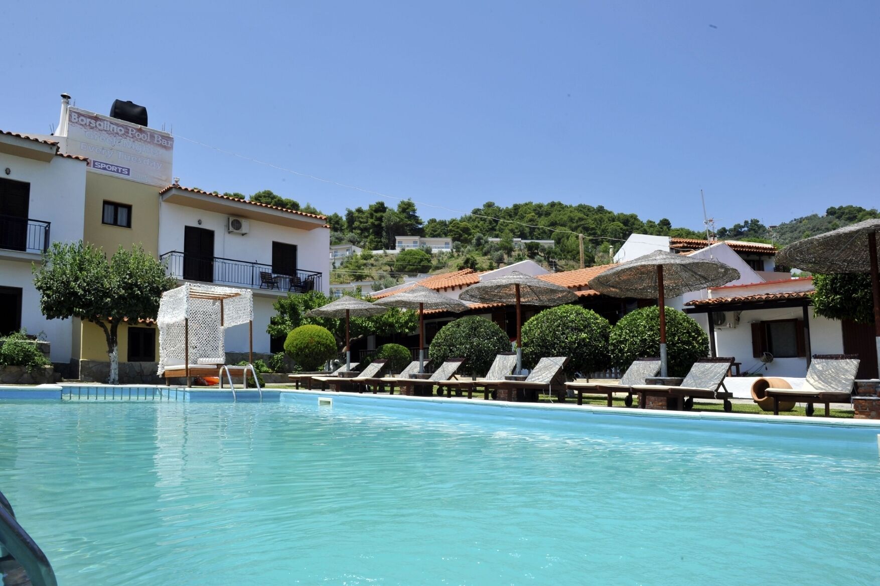 A swimming pool with lounge chairs and umbrellas, a building with a balcony, and a hill with houses in the background.