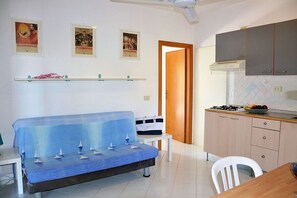 A compact living space with a sofa covered in a blue cloth, a small table with a bag, a kitchenette with cabinets, and a door leading to another room. - Pozzo al Moro Village (Campo nell'Elba)