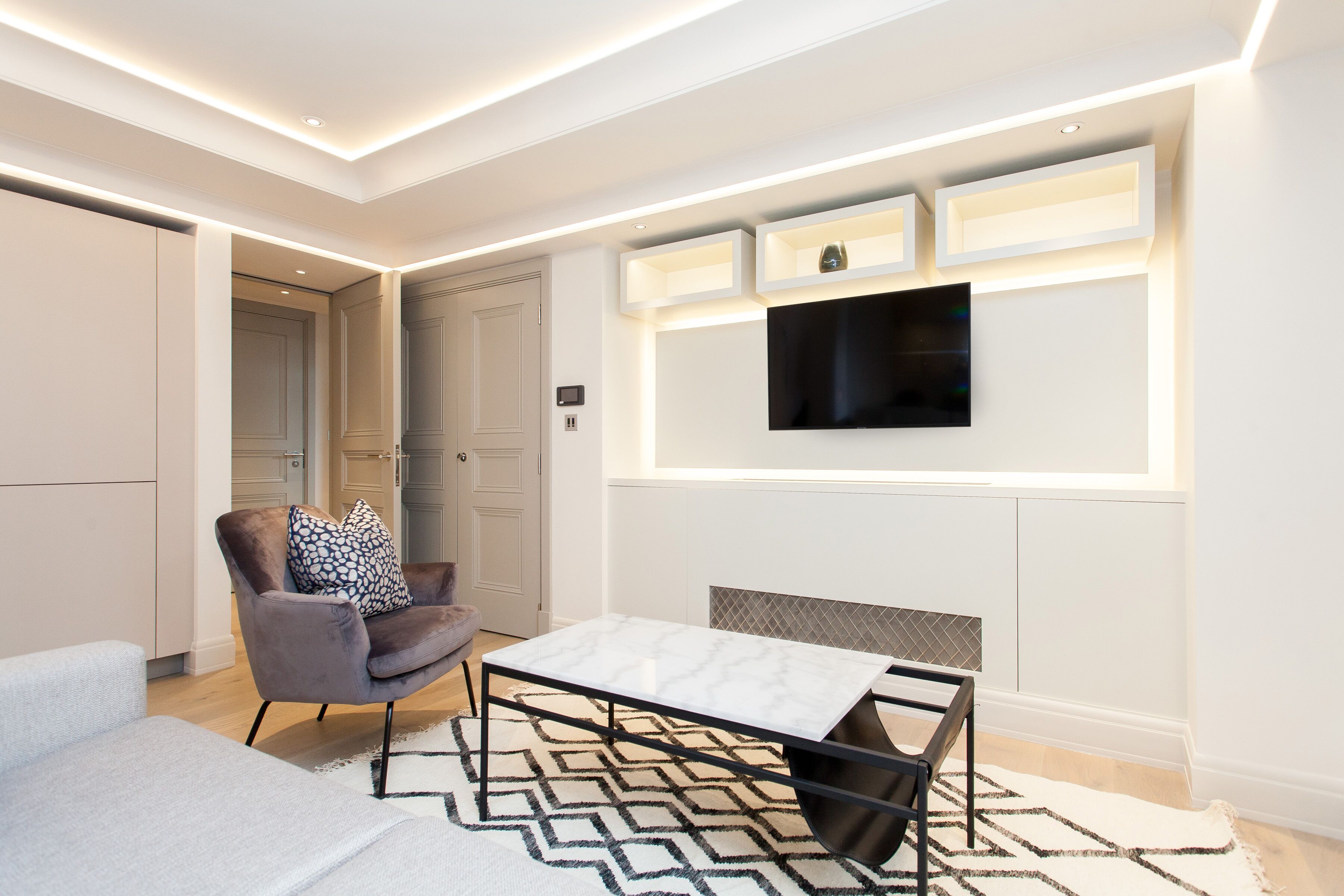 A modern living room with a sofa, a coffee table, and a chair. There is a flat-screen TV mounted on the wall and built-in shelves above it.